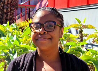 Black woman with neatly braided hair, glasses, cardigan, and knit shirt smiling in front of a garden